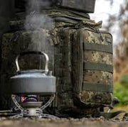 Kettle on a camping stove with a camouflage backpack in the background